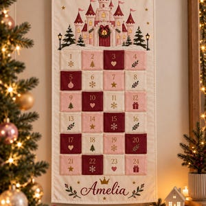 May include: A cream-coloured fabric advent calendar wall hanging featuring a printed pink and red castle at the top. It has twenty-four numbered pockets in alternating shades of pink and red, each with a small holiday icon. The name Amelia is printed in script at the bottom with a gold crown icon.