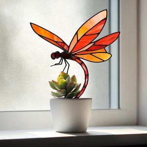 May include: A stained glass dragonfly sculpture in orange and red hues sits above a small succulent plant in a white ceramic pot. The dragonfly has detailed wings and a curved body, creating a decorative accent.