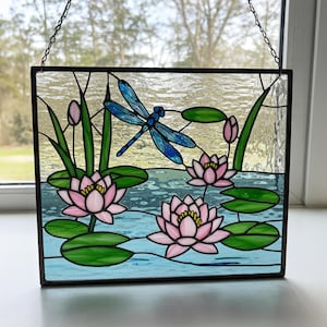 Pode incluir: Uma janela de vitral representando uma libélula azul voando sobre três lírios-d'água cor-de-rosa com folhas verdes de lírios-d'água em um lago azul.