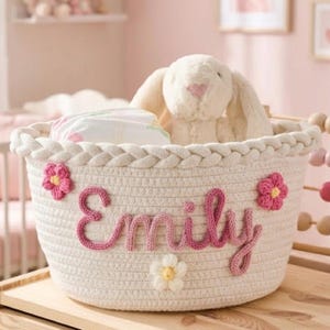 May include: A cream-colored woven basket with the name "Emily" in pink script. The basket is decorated with pink and white flower appliqués and contains a stuffed bunny and other items. The basket is on a wooden surface.