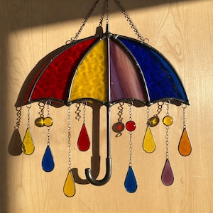 Könnte beinhalten: Bunter Glas-Sonnenschutz in Form eines Regenschirms mit roten, gelben, violetten und blauen Paneelen. Der Schirm hat einen schwarzen Griff und ist mit hängenden tropfen- und runden Glasperlen in verschiedenen Farben verziert. Aufgehängt an Ketten.
