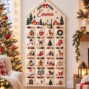 May include: A cream-colored fabric advent calendar with a house-shaped top featuring the name Emma in red script. It has twenty-four numbered pockets decorated with embroidered Christmas motifs like reindeer, snowmen, trees, and houses.