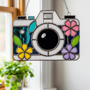 May include: Stained glass camera with floral accents. The camera is white with a black lens and is decorated with pink, yellow, and purple flowers. The camera is hanging from a silver chain.