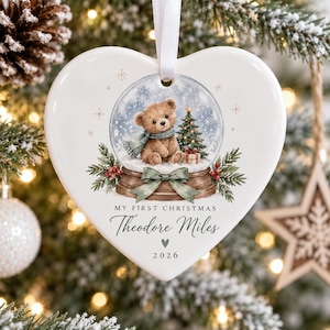 Peut inclure: Un ornement en céramique blanc en forme de cœur illustrant une boule à neige avec un ours en peluche, un sapin de Noël et un nœud vert. Le texte indique My First Christmas, Theodore Miles et 2026. Un ruban blanc est fixé pour suspendre.