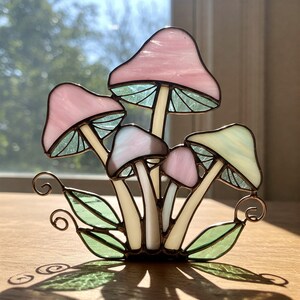Op de afbeelding: Een gebrandschilderde sculptuur van een cluster van paddenstoelen. De paddenstoelenkappen zijn in verschillende tinten roze, bruin en geel, met groene en witte stelen. Groene bladeren en accenten van koperdraad maken het ontwerp compleet.