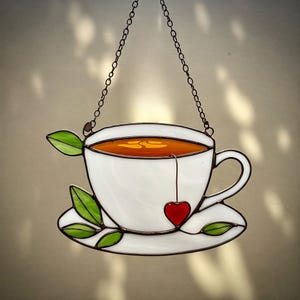 Puede incluir: Pieza de arte en vidriera que representa una taza de té blanca llena de té, una etiqueta de bolsita de té en forma de corazón rojo y hojas verdes. La taza de té está sobre un platillo blanco. Se cuelga de una cadena, ideal para la cocina.
