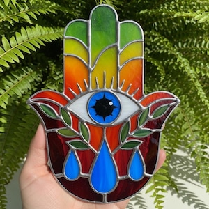 May include: A stained glass Hamsa hand with a vibrant rainbow color scheme. The design features an eye in the center, surrounded by rays and leafy branches. Blue teardrops adorn the bottom. The artwork is set against a backdrop of green foliage.