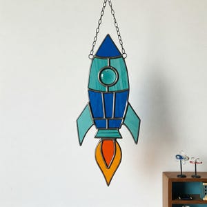 May include: A stained glass rocket ship in shades of blue, teal, and orange, suspended by a chain. The rocket features a flame design at the base and is designed with geometric shapes. A decorative piece for a child's room or space-themed decor.