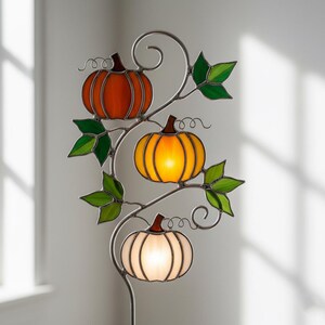 May include: A decorative metal stake featuring three stained-glass pumpkins in orange, yellow, and white. Green leaves and curling vines accent the pumpkins. The pumpkins are illuminated, creating a warm, inviting glow, perfect for fall decor.