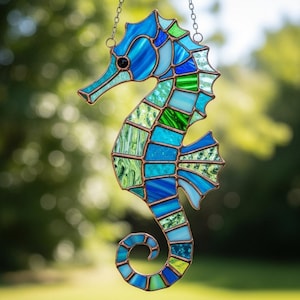 May include: A stained glass seahorse suncatcher in shades of blue and green, with copper-coloured metal framing. The seahorse is suspended by a chain, and the glass pieces create a mosaic effect. The background is a blurred view of green foliage.