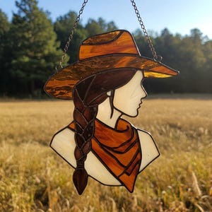 May include: Stained glass artwork depicting a woman in profile wearing a cowboy hat and scarf. The hat and scarf are shades of orange and brown, with a braided hairstyle. The artwork is hanging from a chain.