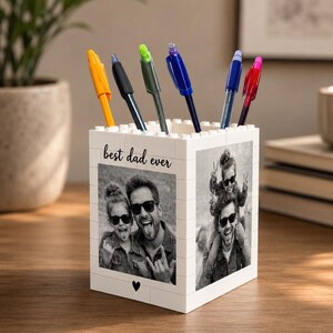 May include: White block-style pen holder with the text "best dad ever" and a heart. The pen holder features black and white photos of a father and child. Several pens in various colours are in the holder.