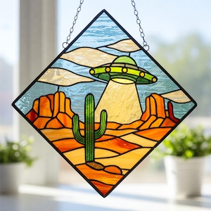 May include: A diamond-shaped stained glass window hanging with a depiction of a desert landscape. The window features a green cactus and a flying saucer hovering over the scene. The sky is blue with white clouds.