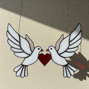 May include: Two white doves holding a red heart in their beaks. The doves are made of stained glass and are hanging from a silver chain.