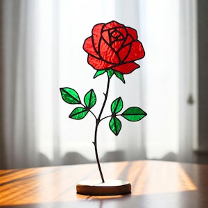 May include: A stained glass rose sculpture featuring a vibrant red flower with green leaves and a dark stem. The rose is mounted on a small, round wooden base, creating a decorative accent piece. The design is reminiscent of a classic rose.