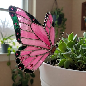 May include: A decorative stained glass butterfly in shades of pink and green. The butterfly features pink wings with green and black detailing, and is embellished with pink jewels. It is placed near a white plant pot with green foliage.
