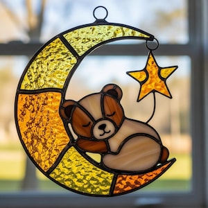 May include: A stained glass suncatcher depicting a sleeping teddy bear on a crescent moon with a star. The moon and star are yellow and orange, the bear is brown and cream. Designed to hang, it's a charming nursery decoration.