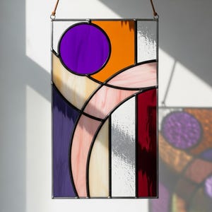 May include: A rectangular stained glass panel with a geometric design. The panel features a large purple circle, orange, pink, white, and red sections, all outlined in black. A leather cord is attached for hanging.