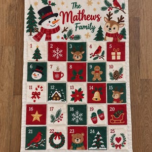 Peut inclure: Un calendrier de l'Avent de Noël en tissu suspendu à une tige en bois avec une corde. Le haut présente un bonhomme de neige, un renne et le texte "The Mathews Family". Il comporte vingt-cinq poches numérotées en rouge, vert et crème, chacune décorée d'icônes de vacances festives comme des arbres, des flocons de neige et des animaux.