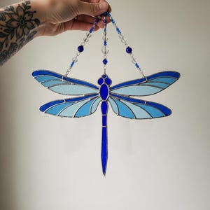 May include: A stained glass dragonfly in shades of blue, with a dark blue outline. The wings are segmented with lighter blue panels. The dragonfly is suspended by beaded strands with blue and clear beads.