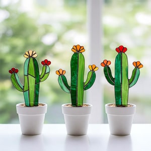 Puede incluir: Tres esculturas de cactus de vidrieras en macetas blancas. Los cactus son verdes con flores coloridas en tonos de rojo, naranja y amarillo. El fondo es una vista borrosa de vegetación y una ventana.