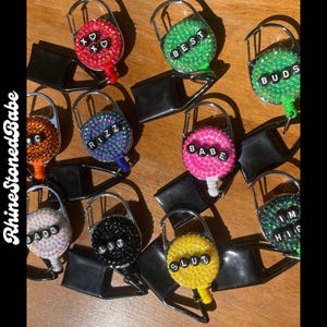 Puede incluir: Una colección de carretes de insignia adornados con pedrería en varios colores, incluyendo rojo, verde, azul, rosa, blanco, negro y amarillo. Cada carrete presenta una palabra o frase diferente escrita con cuentas negras. Los carretes están sujetos a clips negros.