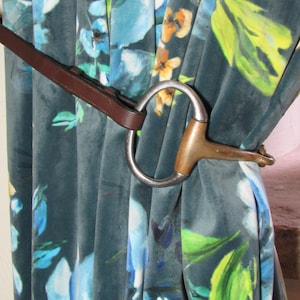 May include: A teal floral curtain held back with a horse bit. The curtain features a watercolour design in blue, yellow, and green. The horse bit is silver and brown, used as a decorative tieback. The curtain is made of a soft, velvety material.