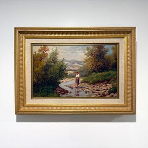 May include: An oil painting in a decorative gold frame depicting a landscape with a stream, green trees, and rolling hills. A figure wearing a red head covering and a dark skirt stands in the shallow water. The scene features natural light.