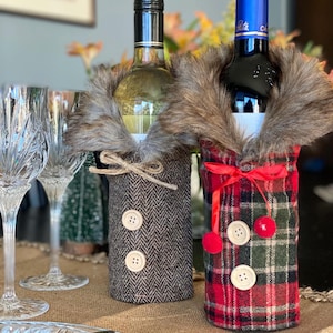 May include: Two wine bottles dressed in festive winter-themed fabric covers. One bottle is wrapped in a brown tweed fabric with two buttons, and the other is wrapped in a red and green plaid fabric with a red pom-pom and two buttons.