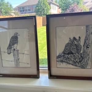 May include: Two framed black and white ink drawings featuring wildlife. The left print depicts a hawk perched on a wooden fence post with barbed wire. The right print shows two owls nestled together on a tree branch. Both have dark wood frames.