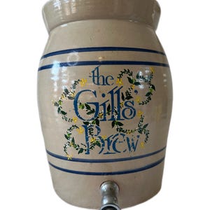以下が含まれることがあります： 青いストライプと「the Gills Brew」の文字が青い筆記体で描かれ、花のアクセントで囲まれた、タン色のセラミック製飲料ディスペンサー。下部に銀色の蛇口が付いています。ヴィンテージスタイルの容器。