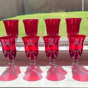 May include: A collection of twelve ruby red wine glasses. The glasses have a classic design with a clear stem and a red band around the middle. The glasses are arranged in two rows of six, with their shadows cast on the surface.