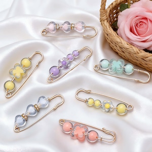 Peut inclure: Plusieurs épingles de sûreté décoratives avec des perles colorées et des motifs floraux ou en forme de cœur. Les épingles sont en métal doré et comportent des perles de différentes couleurs pastel, notamment rose, jaune, violet et transparent. Sur une surface en satin blanc.