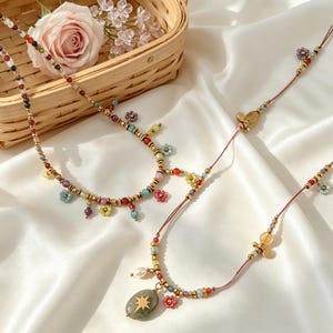 Puede incluir: Dos collares con cuentas multicolores, pequeños dijes en forma de flor y espaciadores metálicos. Un collar incluye un dije ovalado gris con un emblema de estrella dorada y una pequeña perla blanca. Las cuentas están en un cordón fino.