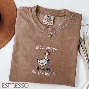 Peut inclure: T-shirt couleur expresso avec l'inscription brod&eacute;e "nicu goose on the loose" et une oie sur un scooter. Le t-shirt a un col rond et des manches courtes. Un collier argent&eacute; avec un pendentif est drap&eacute; sur le t-shirt.