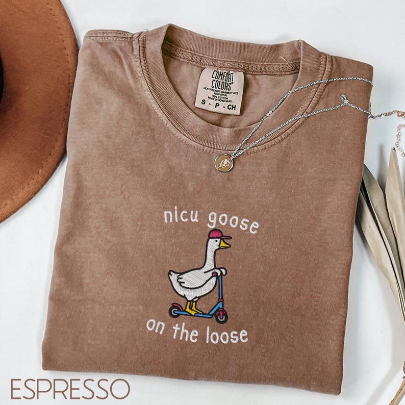 Peut inclure: T-shirt couleur expresso avec l'inscription brod&eacute;e "nicu goose on the loose" et une oie sur un scooter. Le t-shirt a un col rond et des manches courtes. Un collier argent&eacute; avec un pendentif est drap&eacute; sur le t-shirt.