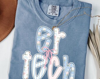 May include: A blue jean colored t-shirt with the words "ER TECH" in a decorative font. The letters are outlined in white and filled with patterns of bows, stripes, and polka dots. A pink bow is tied between the letters. The brand tag "Comfort Colors" is visible.