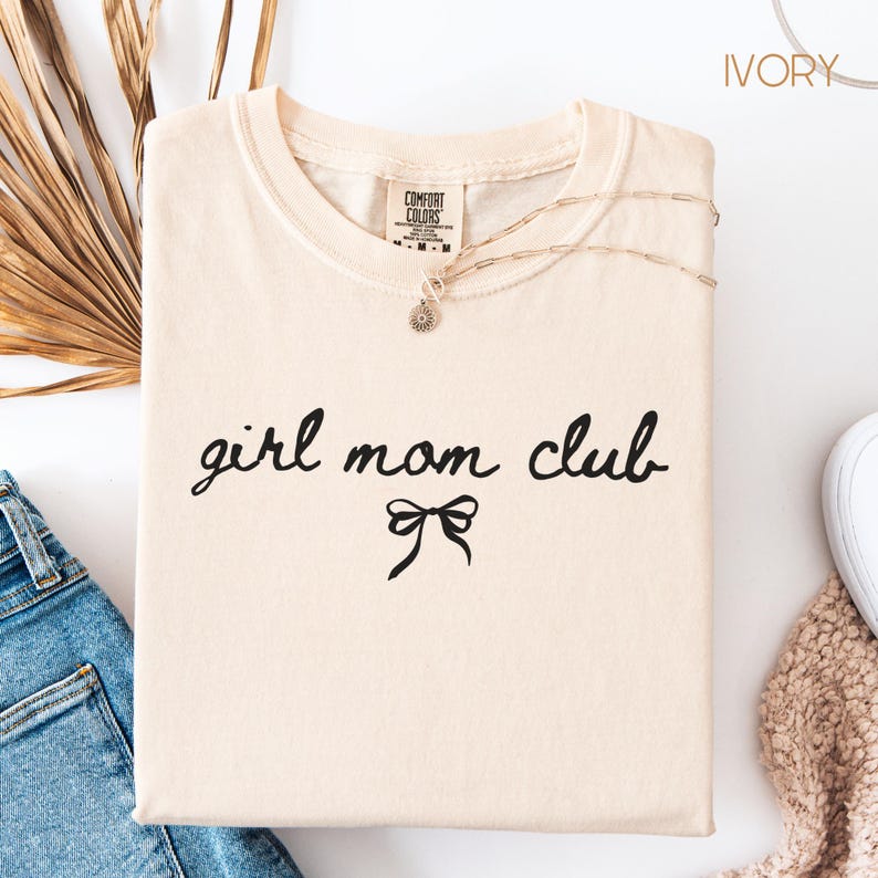 Peut inclure: T-shirt ivoire avec le texte "girl mom club" en &eacute;criture noire avec un n&oelig;ud noir en dessous.