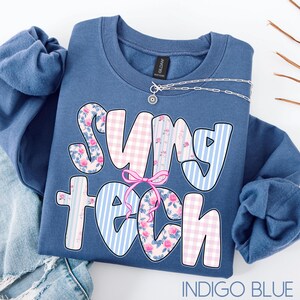 May include: Indigo blue sweatshirt with the text "SUNY TECH" in a playful font. The letters feature floral, gingham, and striped patterns. A silver necklace with a small pendant is draped across the top. The sweatshirt is folded on a white surface.