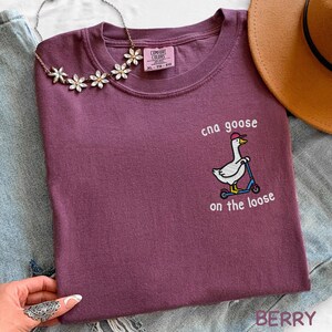 Op de afbeelding: Bessenkleurig T-shirt met de geborduurde tekst "cna goose on the loose" en een gans op een scooter. Het shirt heeft een ronde hals en een Comfort Colors label.