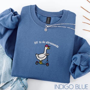 May include: Indigo blue sweatshirt featuring the embroidered text "Off to do ultrasounds" and a goose on a scooter, wearing a red cap. A silver necklace with a sun pendant is draped across the sweatshirt. The sweatshirt is a shade of indigo blue.