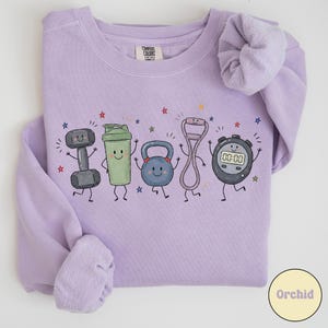 Könnte beinhalten: Ein hellviolettes Sweatshirt mit Rundhalsausschnitt und einem Grafikdruck von fünf anthropomorphen Fitnessartikeln, darunter eine Hantel, ein Shaker, eine Kettlebell, ein Widerstandsband und eine digitale Stoppuhr, umgeben von kleinen bunten Sternen.