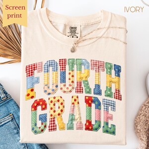 May include: Ivory-colored t-shirt with the words "FOURTH GRADE" in a patchwork design. The letters are in various colors and patterns, including polka dots and gingham. The shirt has a crew neck and a Comfort Colors tag.