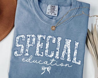 May include: A blue jean colored t-shirt with the words "SPECIAL education" in white floral print. The shirt has a crew neck and short sleeves. A gold necklace with a pendant is draped across the shirt.