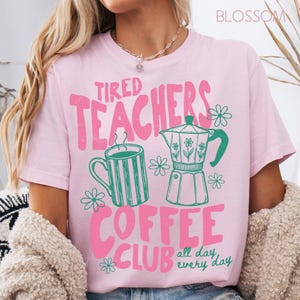 May include: Light pink t-shirt with the text "Tired Teachers Coffee Club" in pink and green. The design includes a coffee mug, a coffee pot, and floral accents. The words "all day, every day" are also included.