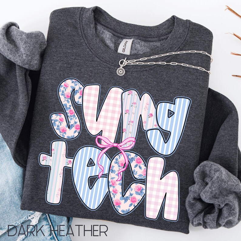 May include: Dark heather gray sweatshirt with the text "SUNY TECH" in a colorful design. The letters feature floral, gingham, and striped patterns in pink and blue, with a pink bow. A silver necklace is draped across the top.