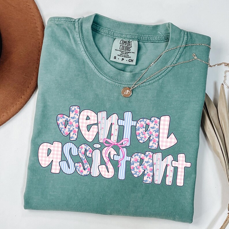 Pode incluir: Uma camiseta Comfort Colors verde-azulado com as palavras "dental assistant" em uma fonte l&uacute;dica. As letras s&atilde;o decoradas com estampas florais, xadrez e listras. Um colar de ouro com um pingente repousa sobre a camiseta.