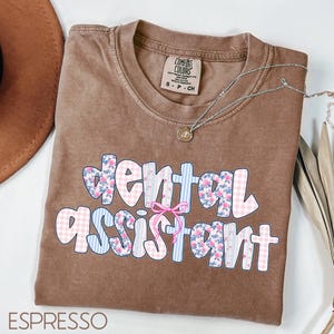 Pode incluir: Camiseta cor de caf&eacute; com as palavras "dental assistant" em um design floral e xadrez. Um colar de prata com um pingente est&aacute; drapeado sobre a camiseta. A camiseta &eacute; da marca Comfort Colors.