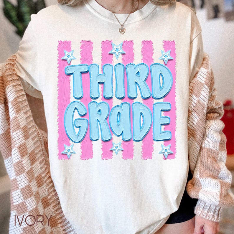 May include: Ivory-colored sweatshirt with the words "THIRD GRADE" in light blue, outlined in a darker blue, set against a pink striped background. Small blue stars are scattered around the text. The sweatshirt has a crew neck.