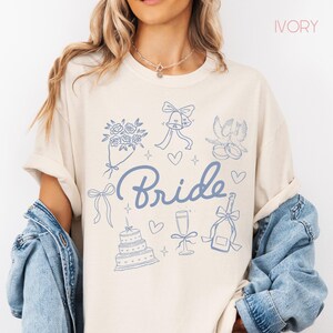 May include: Cream-coloured t-shirt featuring a blue graphic design. The design includes the word "Bride" in cursive, alongside illustrations of flowers, bells, doves, hearts, a cake, champagne, and a bow. The word "IVORY" is printed at the top.
